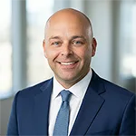 Portrait of Mil Radenkovic in a navy suit, white shirt, and blue tie, standing indoors with blurred background. He is the Sales Manager at Hunt Chrysler Fiat, Milton, Ontario, Canada.