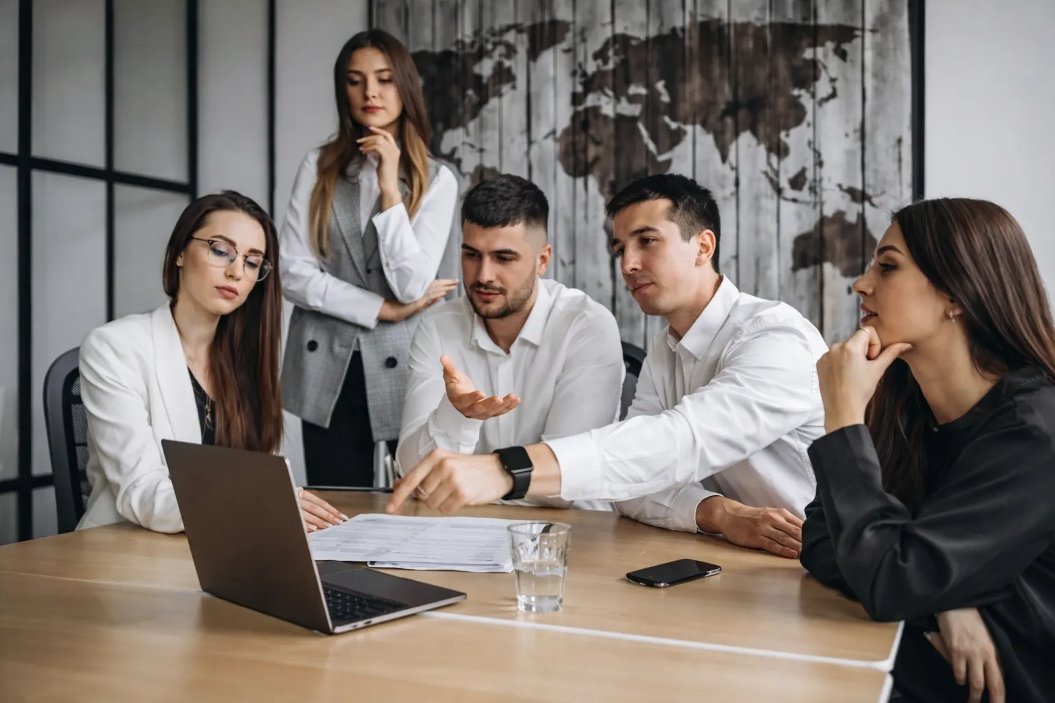 Five business professionals discussing documents and laptop during a meeting in a modern office with a world map on the wall.